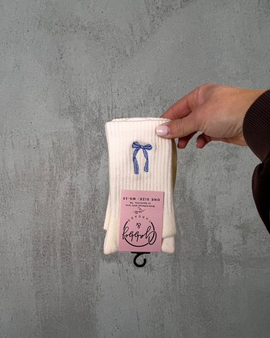 a hand holds up a pair of white grip socks with a blue coquette bow embroidered on it against a grey background
