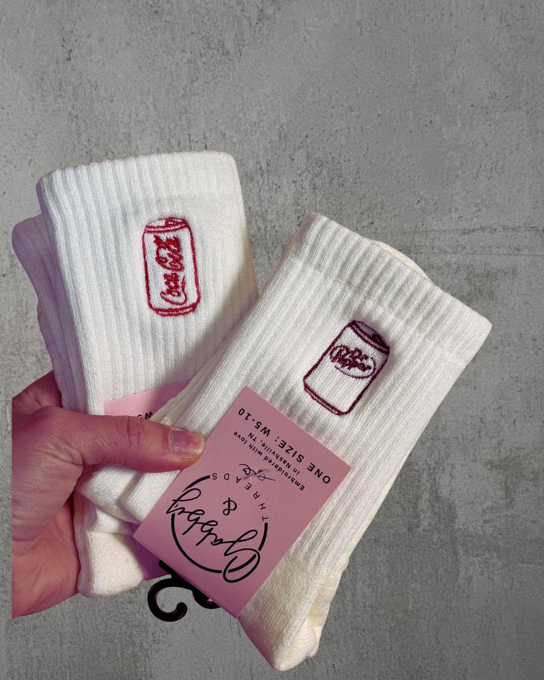 two pairs of white grip socks held up by a hand. one has an embroidered coca cola on them and the other has an embroidered dr pepper can