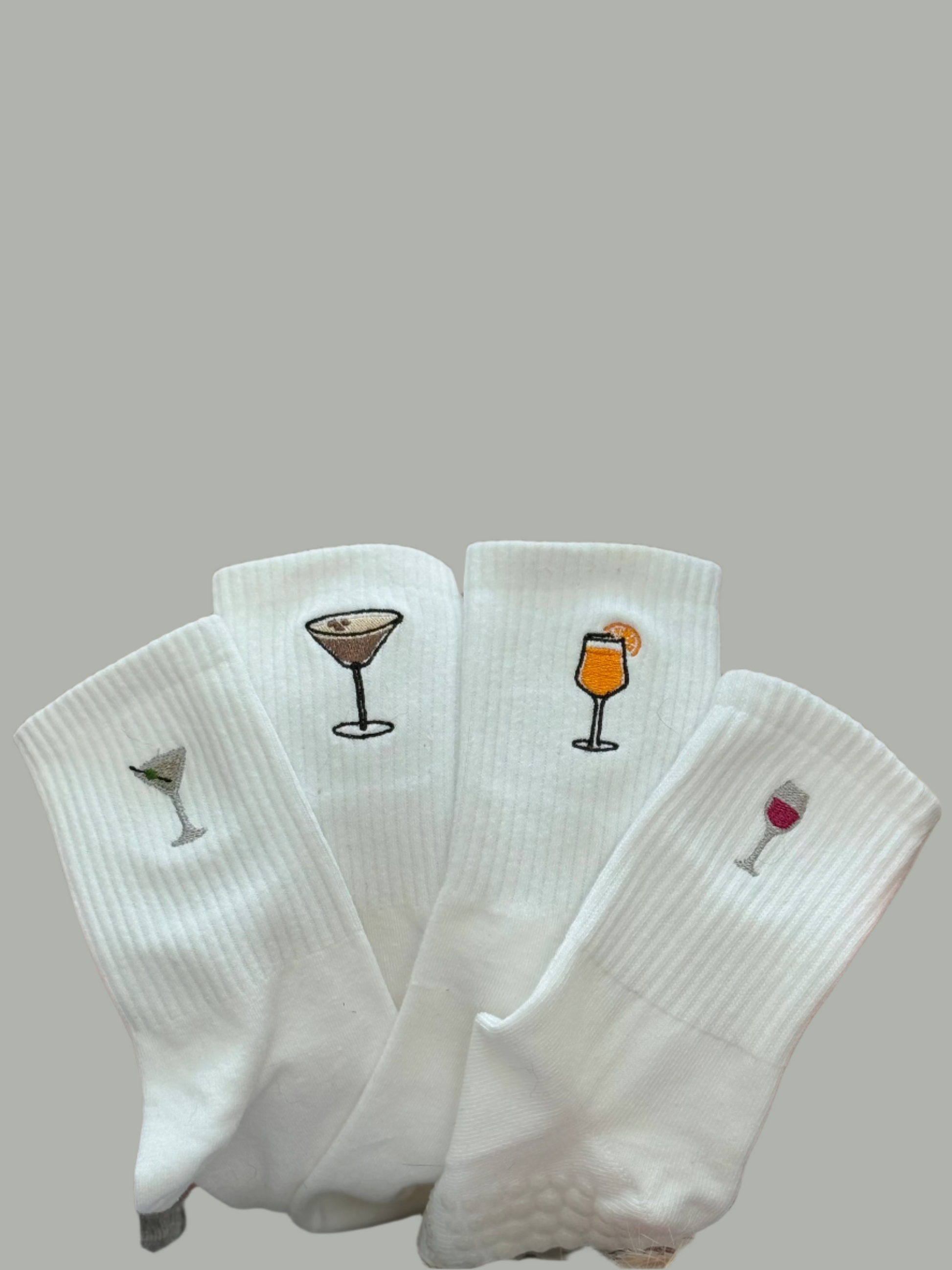 Four different  pairs of white grip socks with different cocktails embroidered on each one. They include espresso martini, dirty martini, aperol spritz and a wine glass.