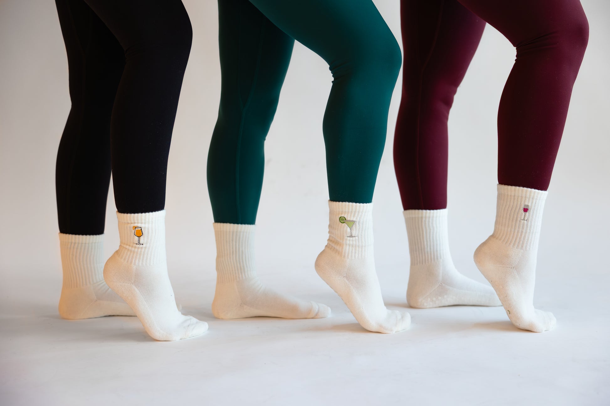 Three pairs of legs wearing different colored leggings and white socks on a light background. each pair of white grip socks has a different cocktail embroidered on it