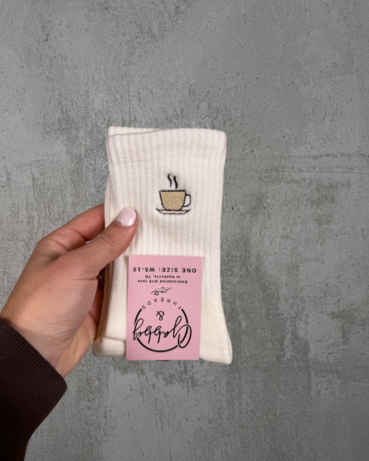 A pair of White grip socks with coffee cup design held by a hand against a gray background