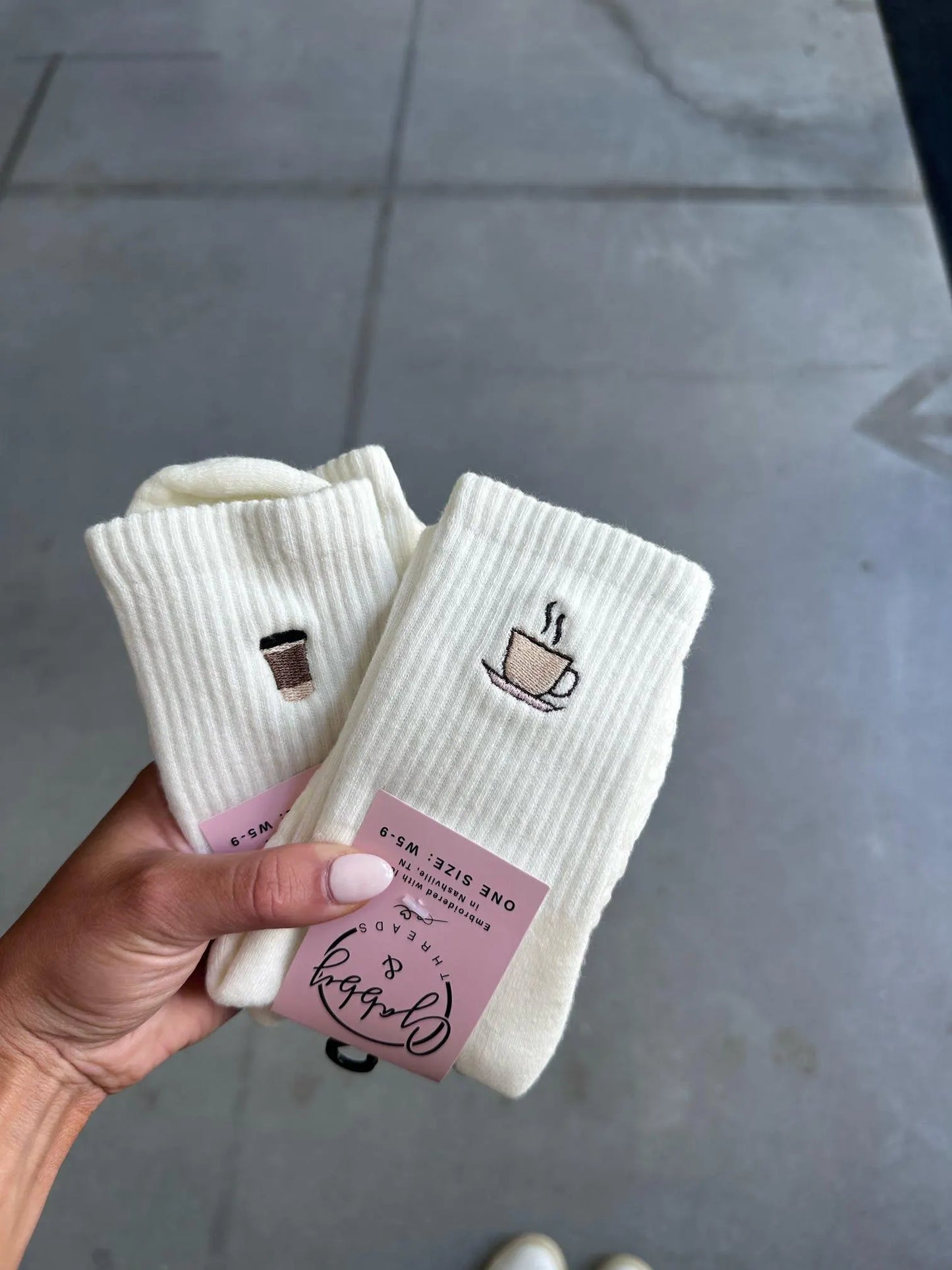 White socks with coffee-themed embroidery held by a hand on a gray floor.