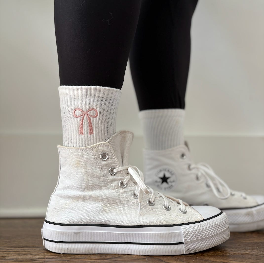 A close up of a woman wearing white high-top converse and everyday crew socks with a light pink coquette bow on them