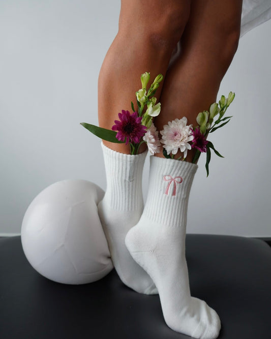White socks featuring a pink coquette bow with floral arrangements held between them on a neutral background