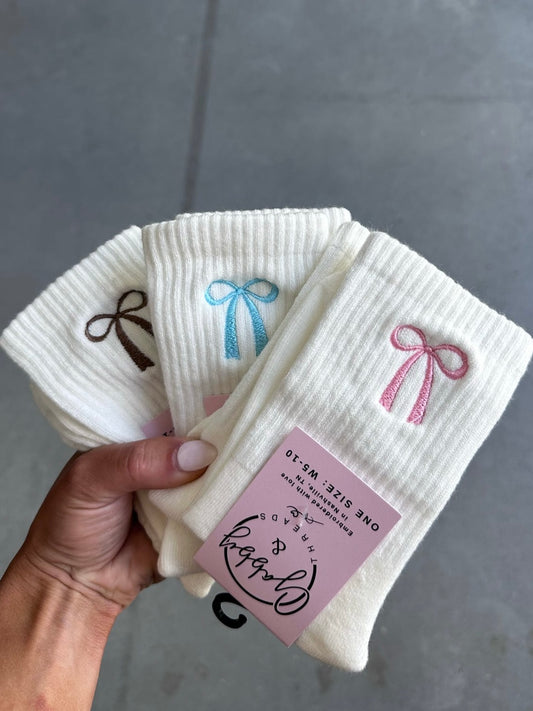 White crew socks with embroidered bow designs held by a hand against a gray background