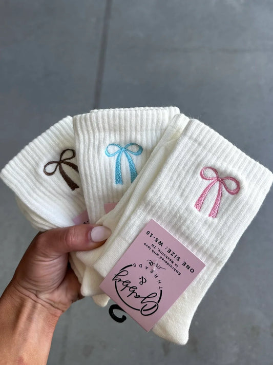 three pairs of white grip socks with different colored embroidered coquette bows on them against a gray background