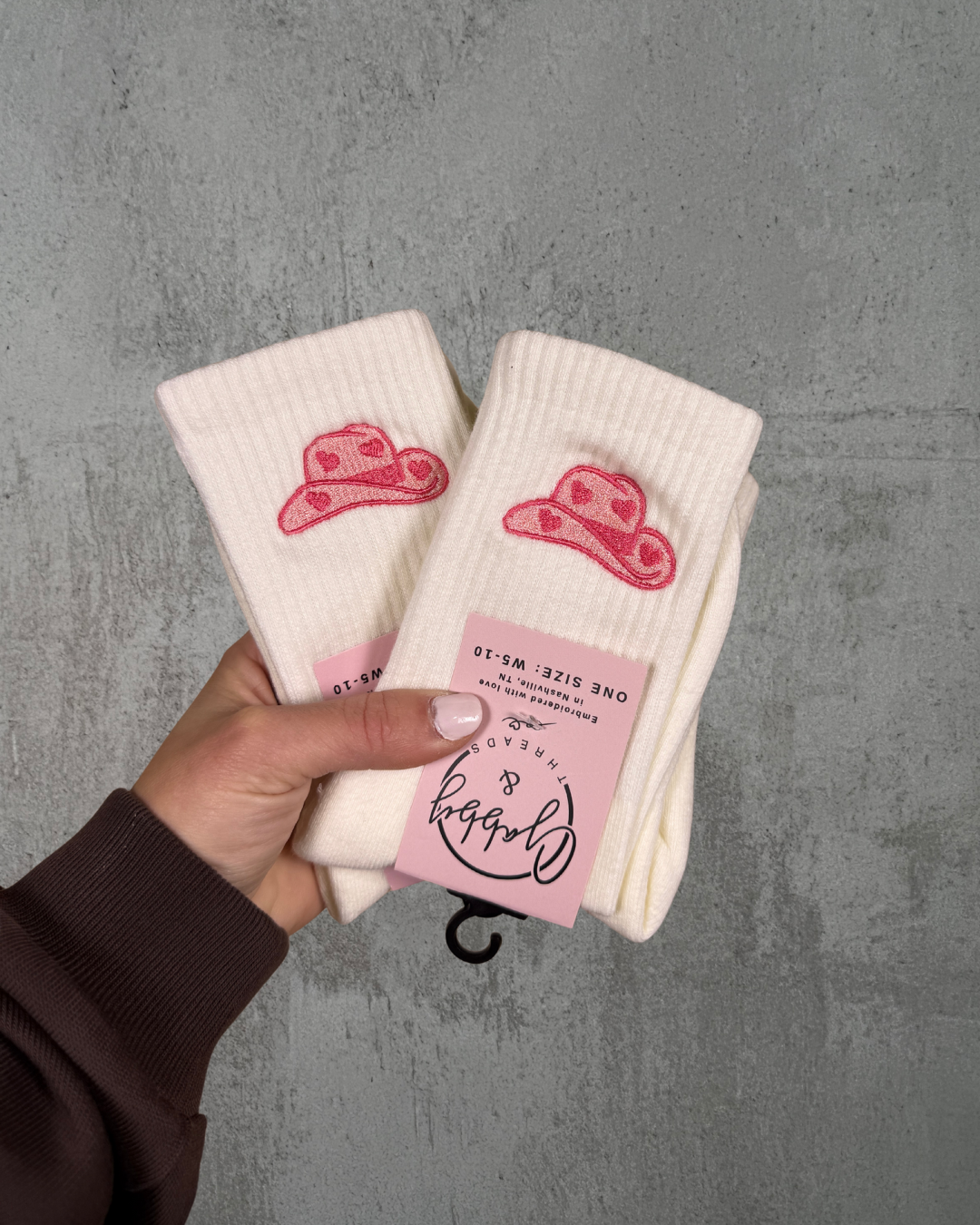 a hand holds up two pairs of socks, with pink embroidered cowgirl hats on them against a gray background
