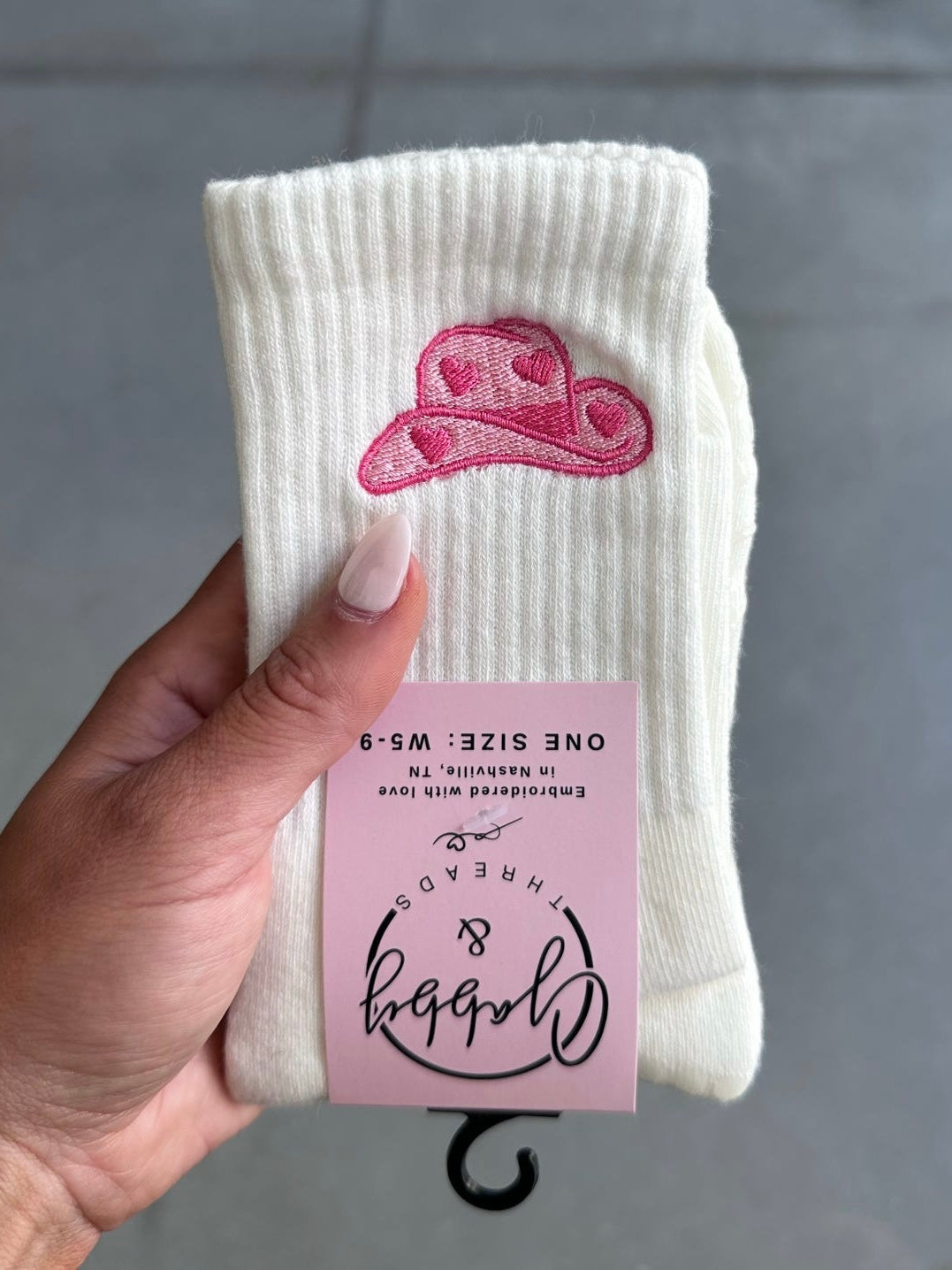White sock with pink cowgirl hat embroidery design held by a hand against a gray background