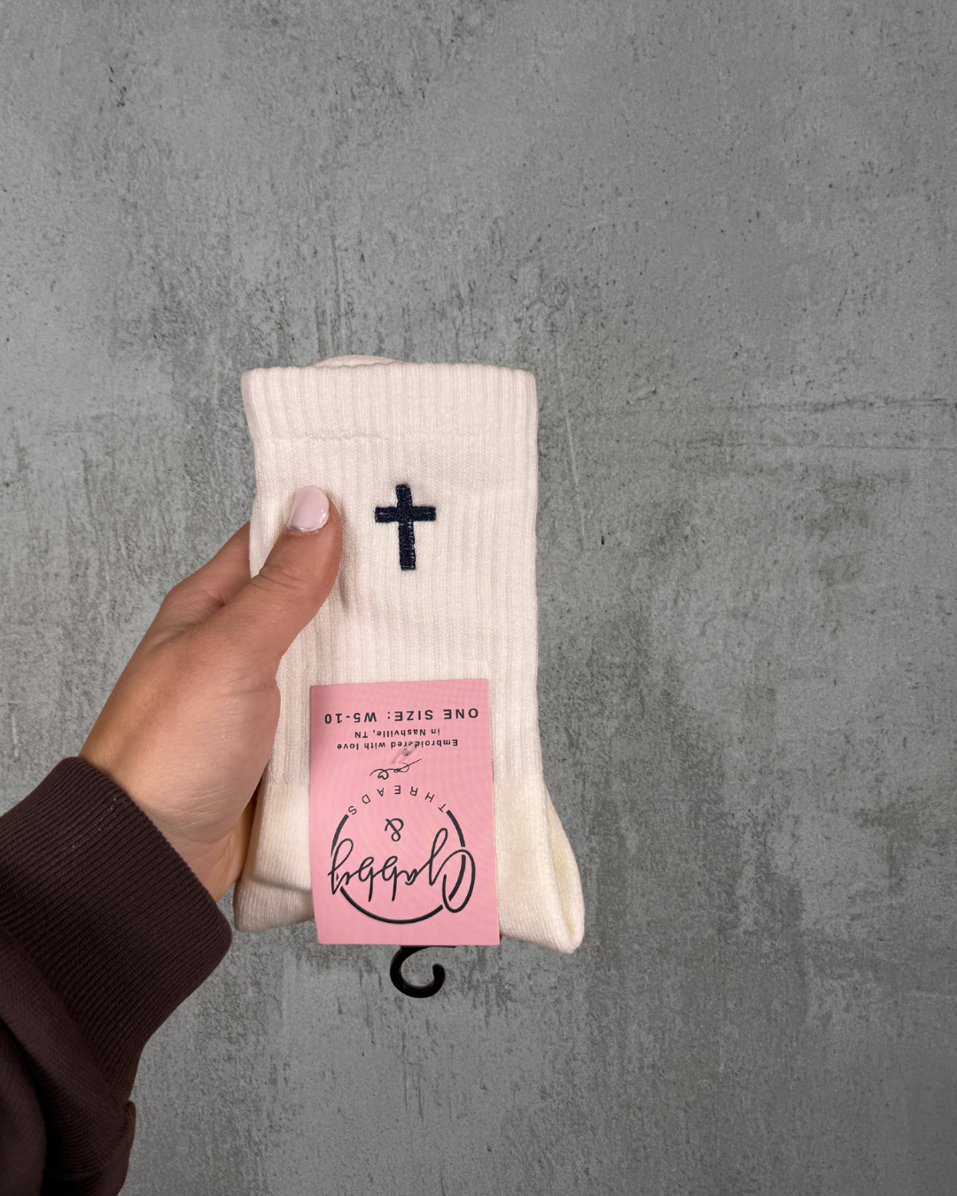 a hand holds up a pair of white grip socks with a cross embroidered on it against a grey background

