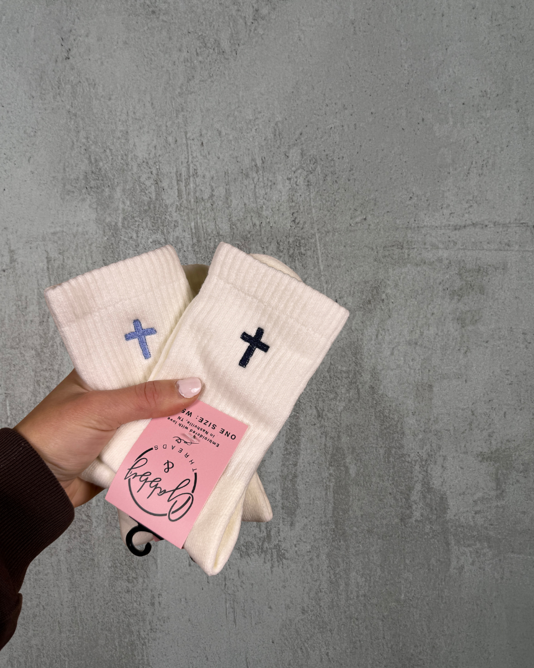 a hand holds up two pairs of socks, both have crosses embroidered on them against a gray background