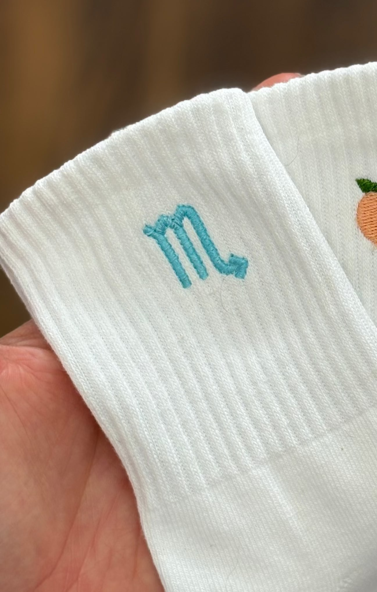 White grip socks with embroidered horoscope held in a hand against a wooden background.