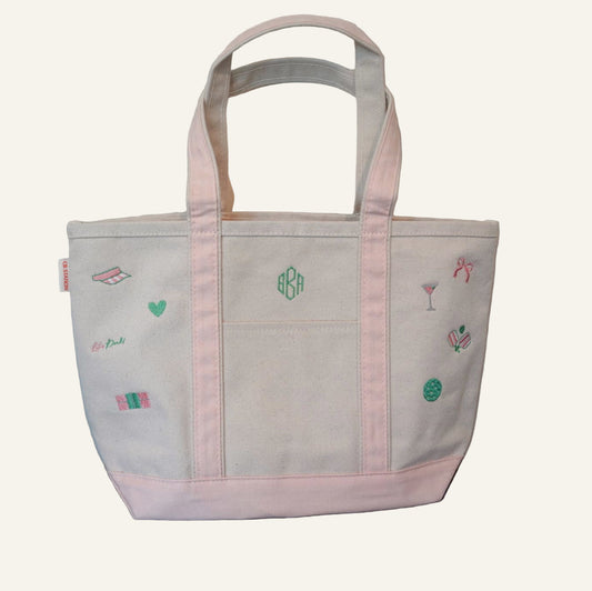 Tan canvas tote bag with light pink straps and embroidered pickleball designs and monogram on a wooden surface.