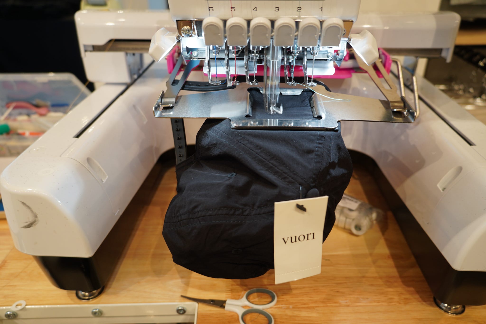 Embroidery machine with a black cap on a wooden table, scissors nearby