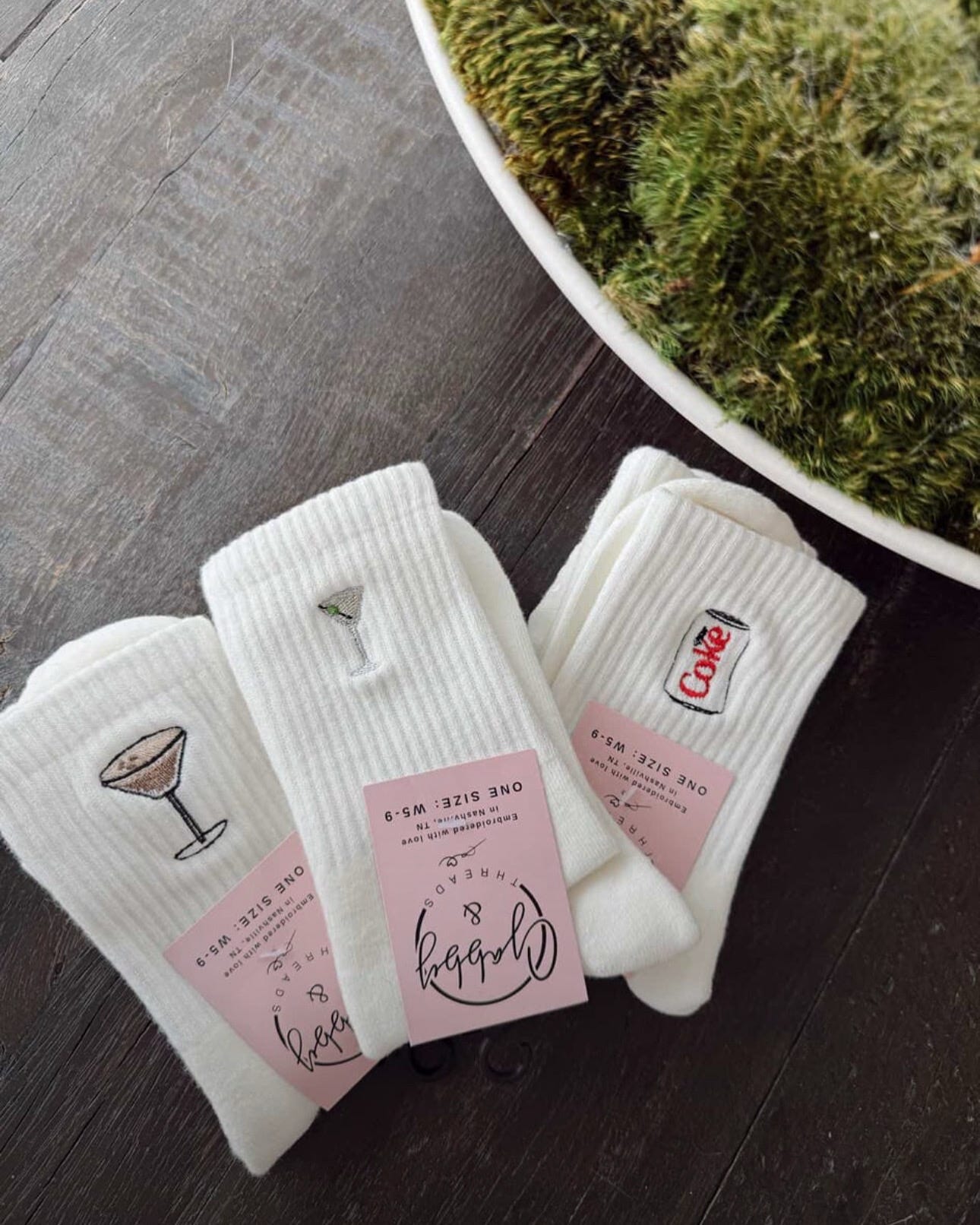 three pairs of white grip socks with different embroidered icons. One has an espresso martini, one has a dirty martini, diet coke