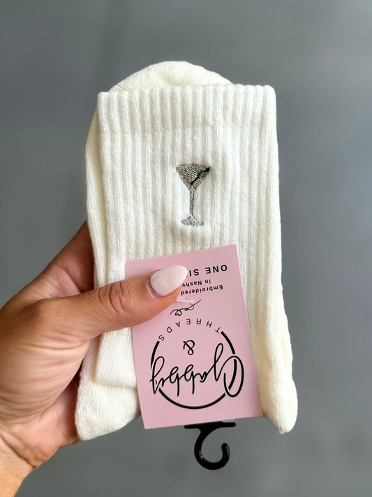White socks with an embroidered dirty martini design held by a hand against a gray background