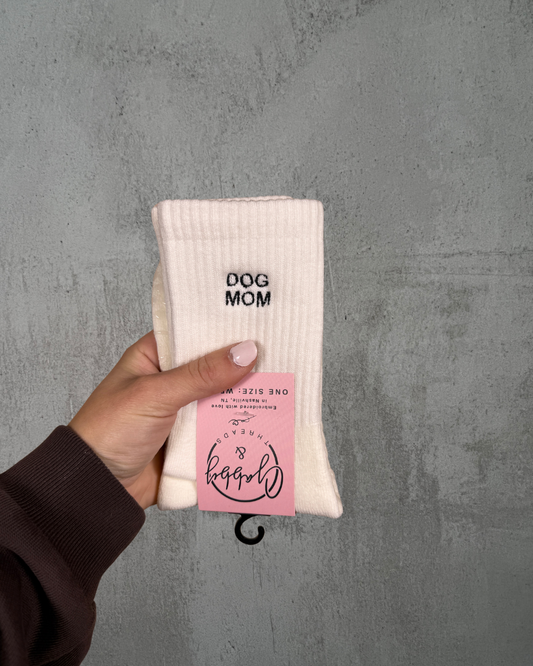 A pair of white socks with the text "dog mom" embroidered on them against a grey background