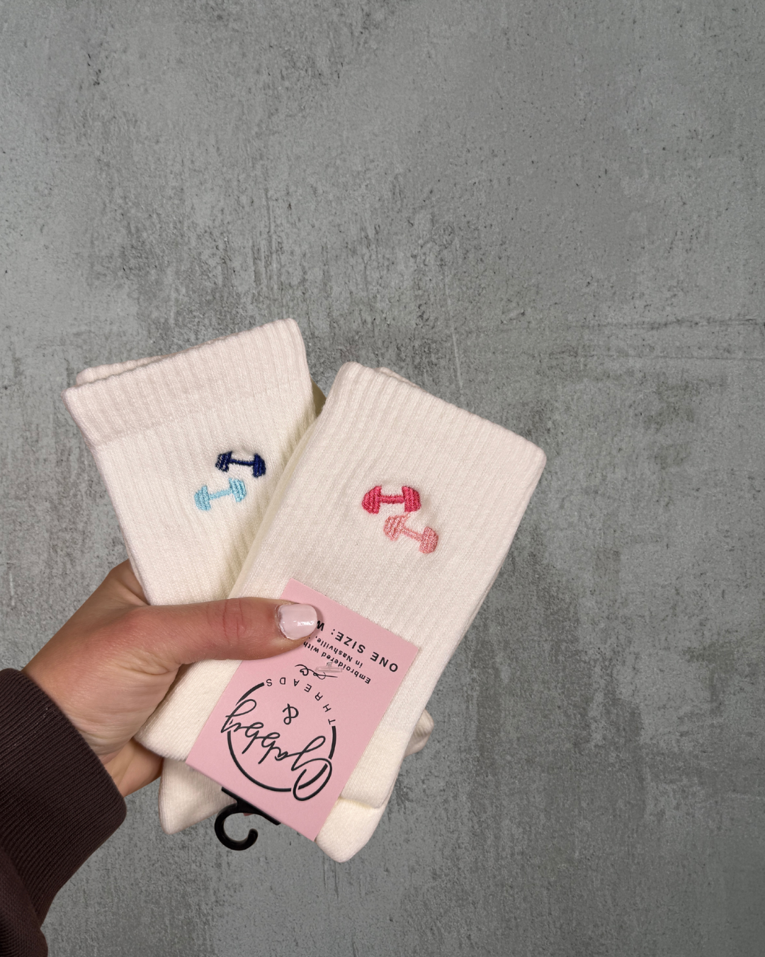 A hand holds two pairs of White grip socks with embroidered dumbbells on them 