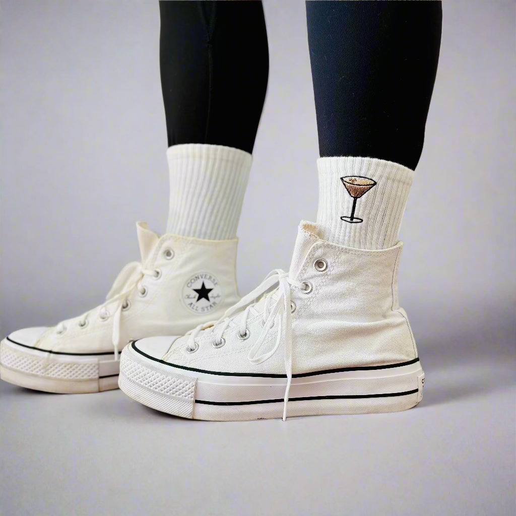 A person is wearing white converse with crew socks that have an embroidered espresso martini detail on them