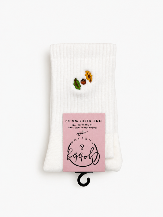 a pair of white socks with two fall inspired leaves and an acorn embroidered on them