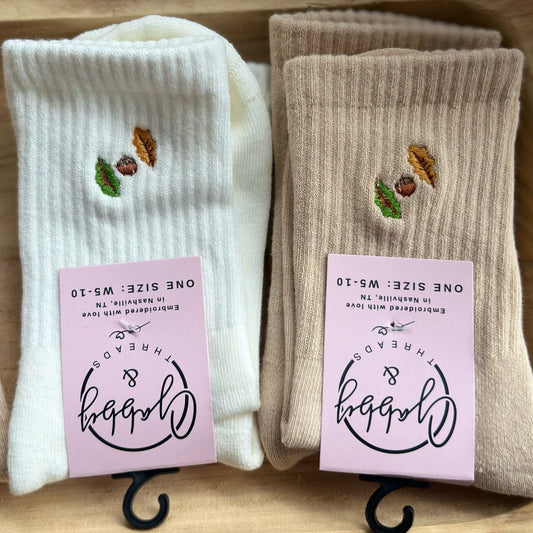 two pairs of grip socks in white and beige with embroidered fall leaves on them