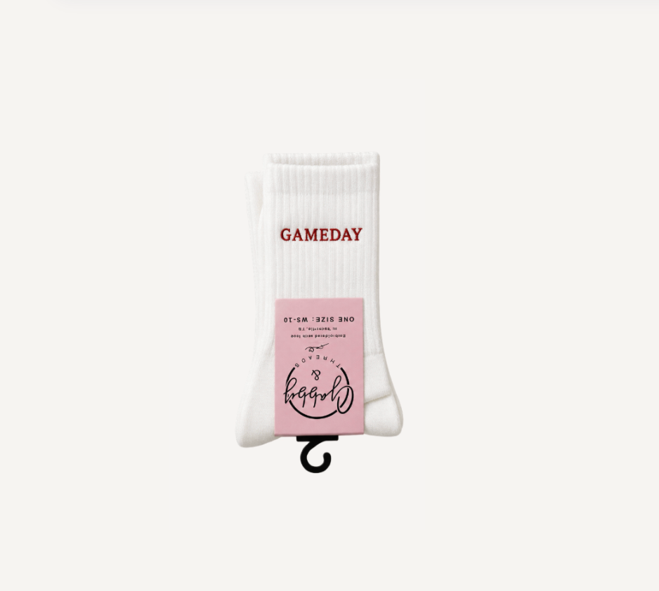 A single white crew sock hanging on a black hook against a plain off-white background. The sock has the word 'GAMEDAY' printed in red near the top and a pink paper label wrapped around the lower half with black text and logo.