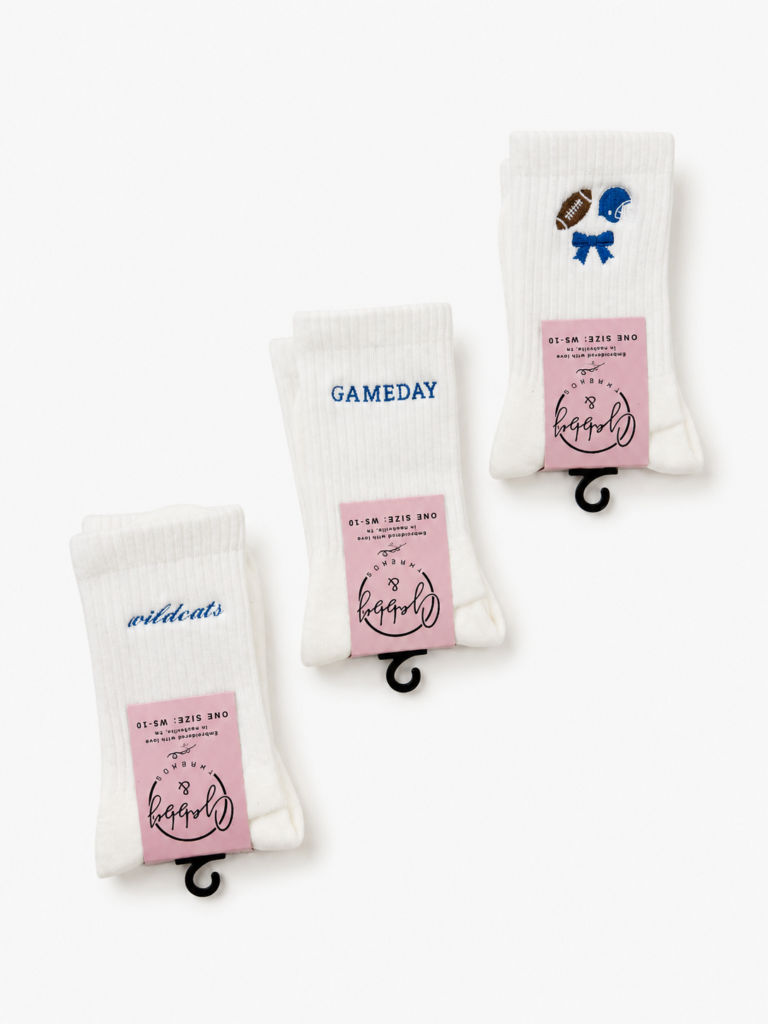 Three pairs of white ribbed socks displayedagainst a clean white background. Each pair has a pink paper label with the brand name gabby and threads and different blue graphic details: one pair has 'wildcats' in blue script, one has 'GAMEDAY' in blue block letters, and one has a blue bow and silver football graphic.