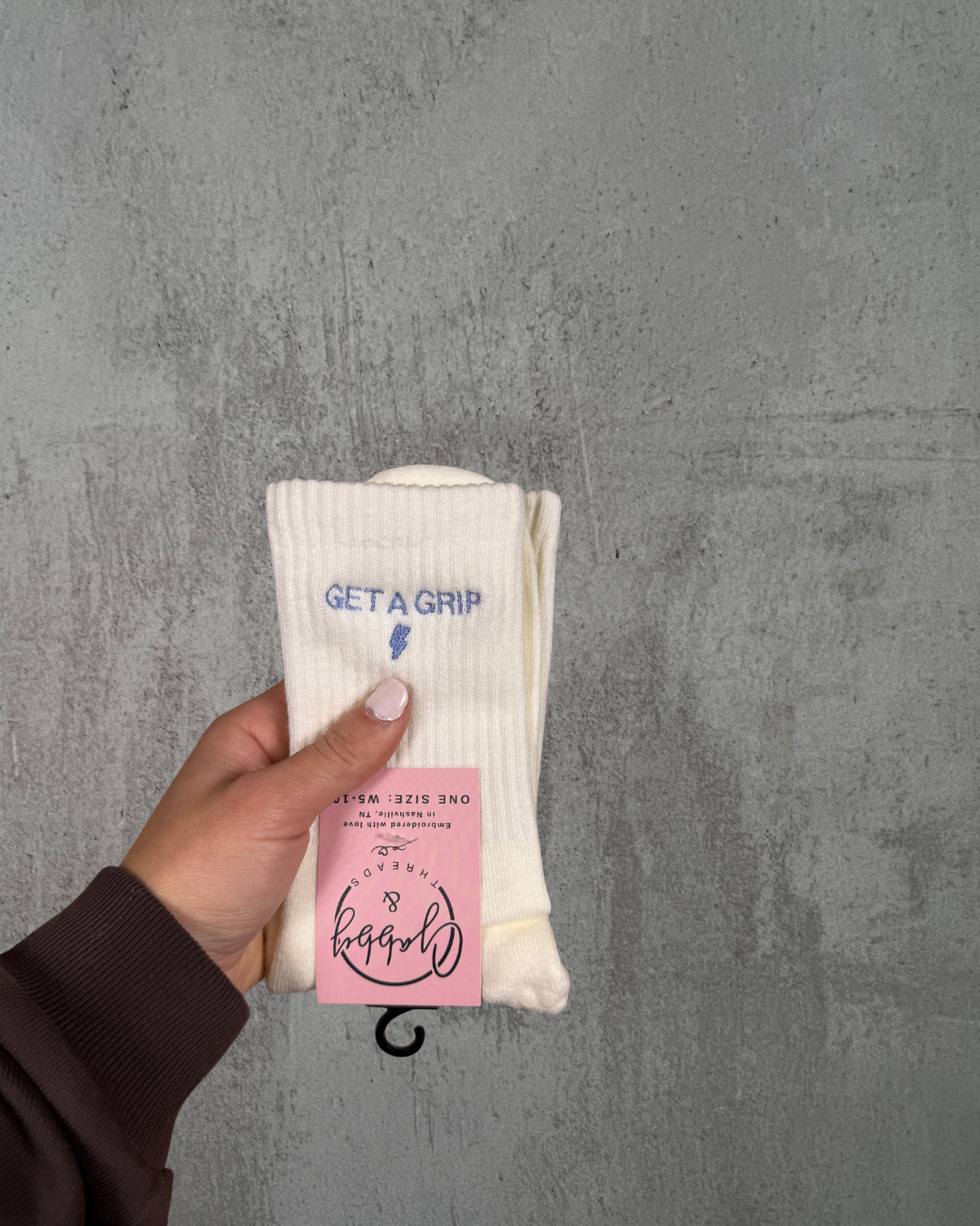 a hand holds up a pair of white grip socks with the text "get a grip" embroidered on it against a grey background