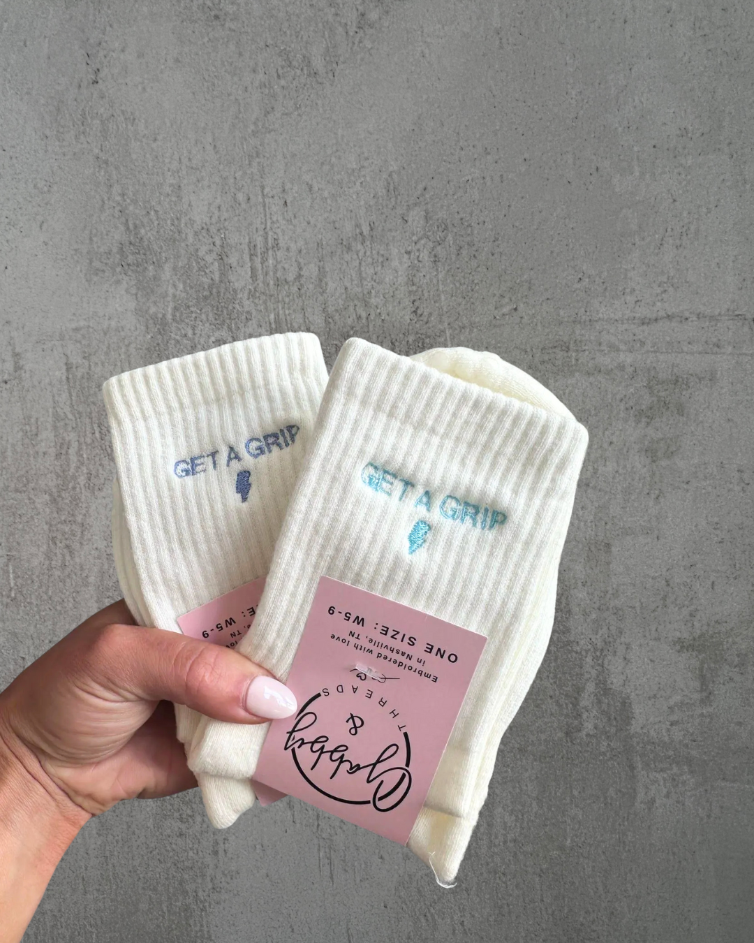 a hand holds up two pairs of white grip socks with the text "get a grip" embroidered on them against a grey background