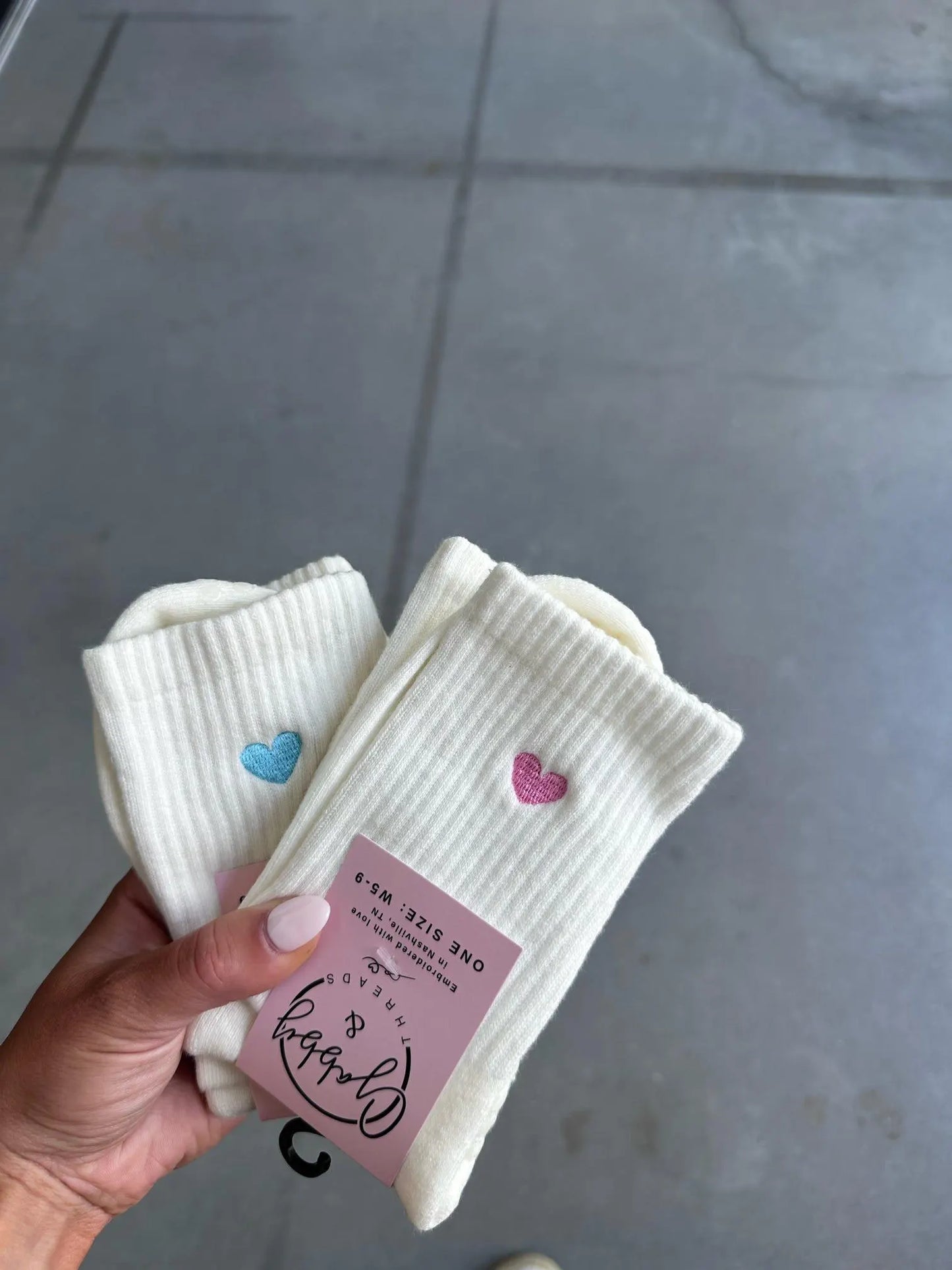 Two pairs of White socks with an embroidered heart held by a hand against a gray background