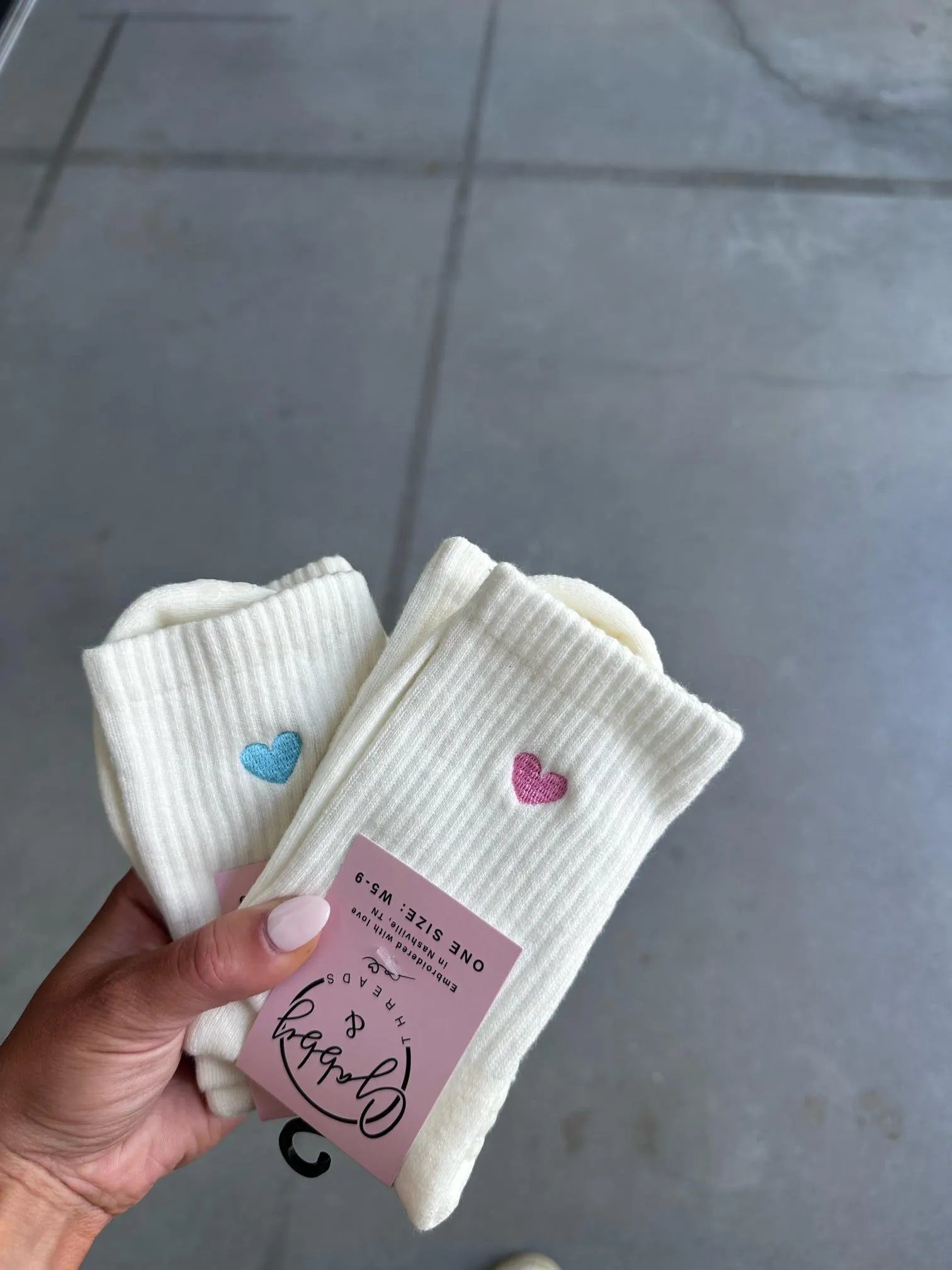 Two pairs of White socks with an embroidered heart held by a hand against a gray background