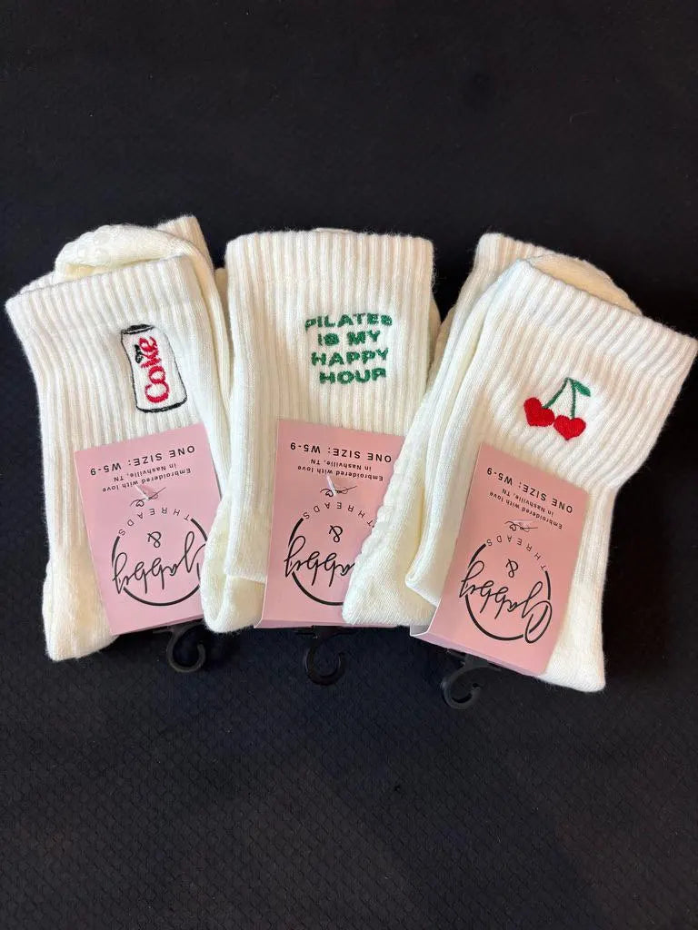 three pairs of white grip socks, all featuring different embroidered designs. One has a diet coke can, one says pilates is my happy hour and the other one has embroidered cherries on a black background