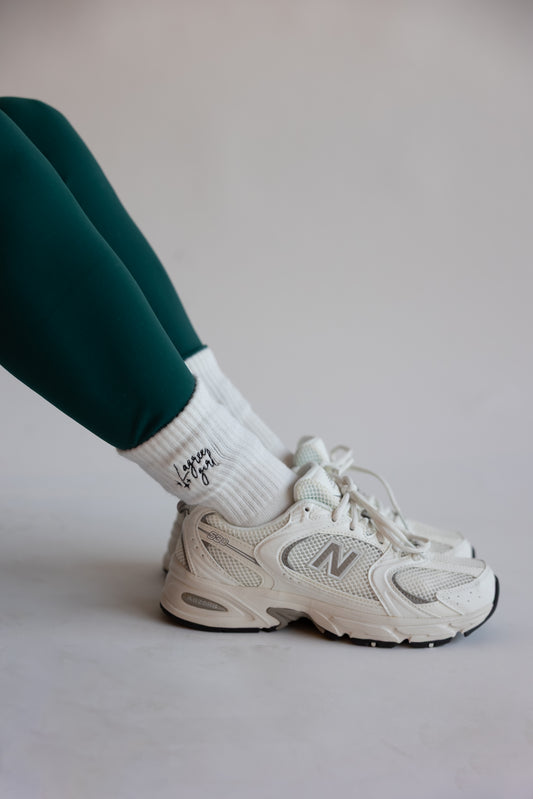 a person wears new balance sneakers, a teal workout set and a pair of white crew socks with the words "Lagree girl" embroidered on them