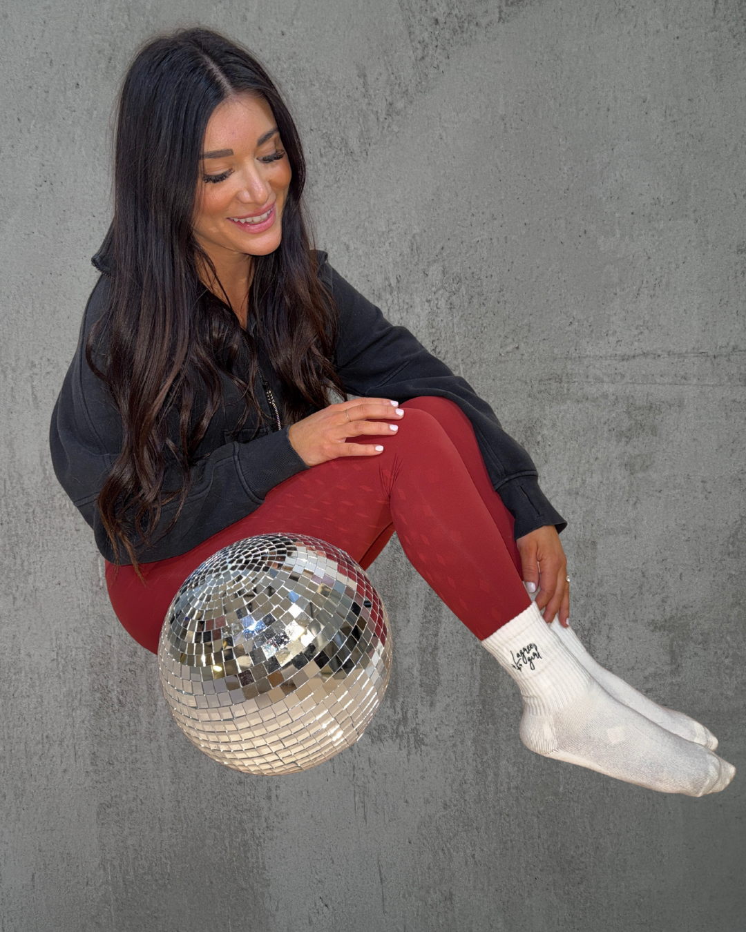 a girl wearing grip socks that say "Lagree Girl" sits next to a discoball against a grey background