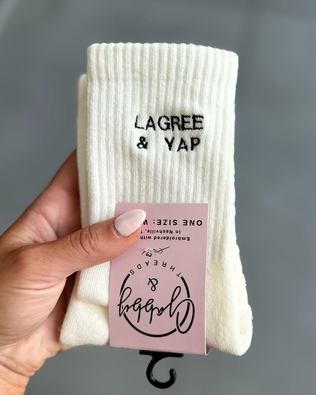White socks with 'Lagree & Yap' branding held by a hand against a gray background