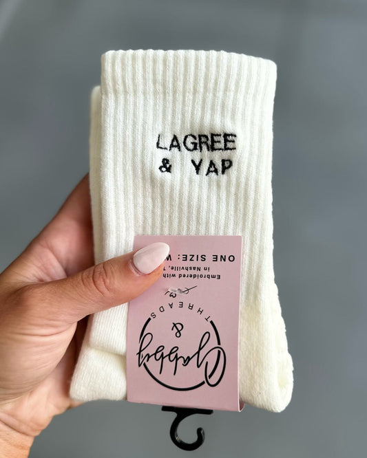 White socks with 'Lagree & Yap' branding held by a hand against a gray background