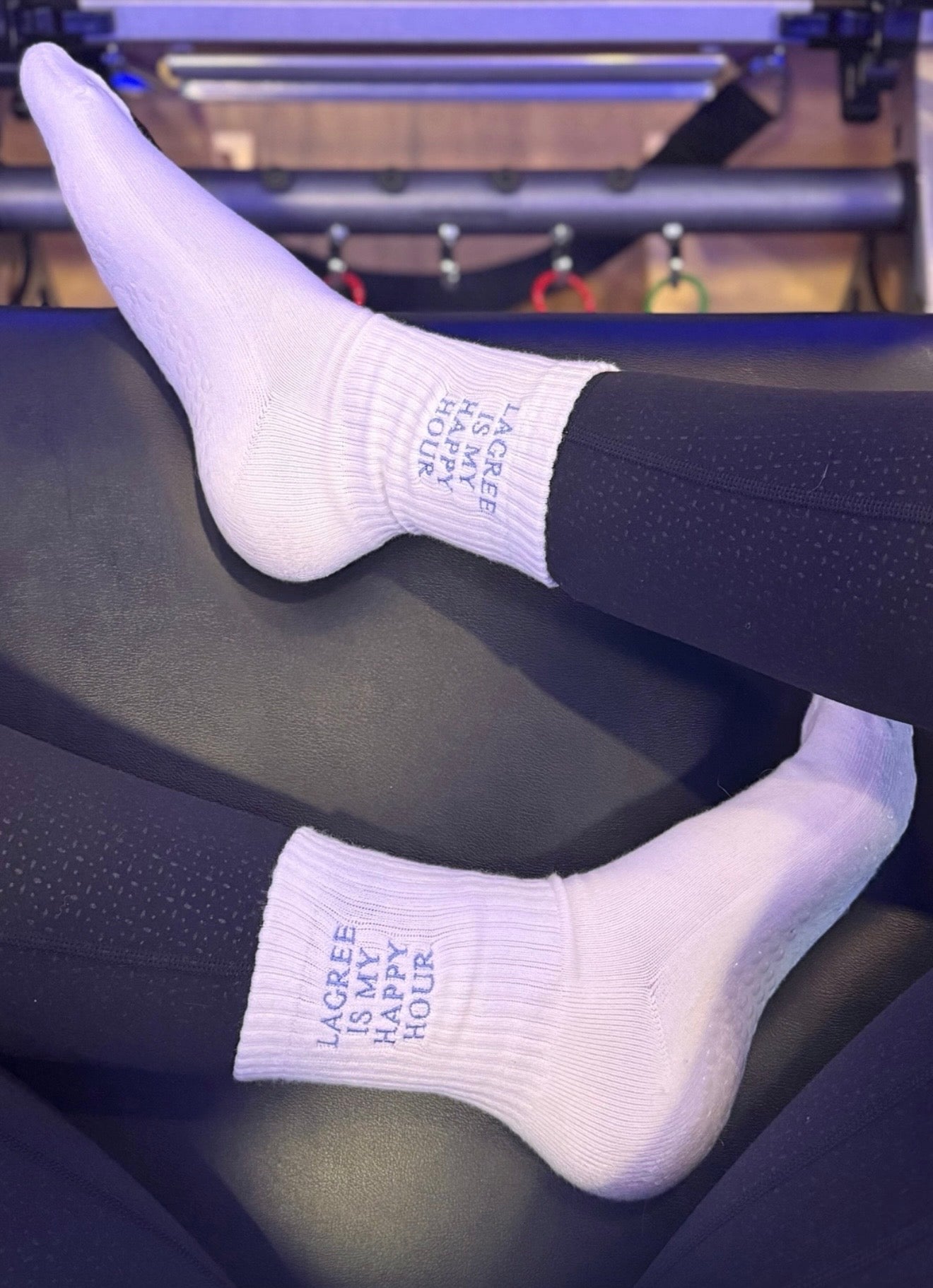 a pair of white grip socks are on someone's feet that say "lagree is my happy hour" while sitting on a pilates reformer 