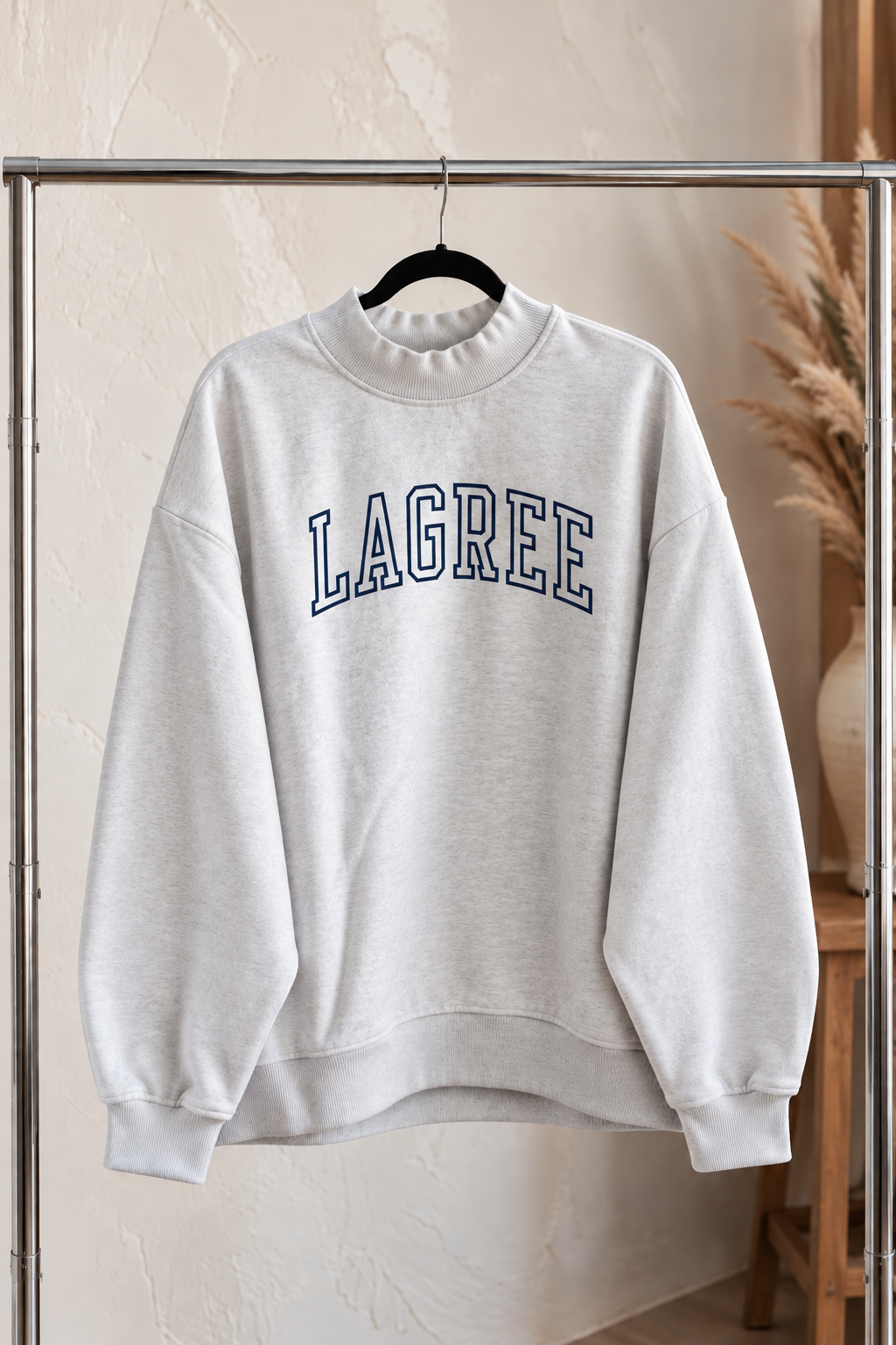 a heather gray mock neck sweatshirt is hanging on a clothing rack. The sweatshirt is embroidered with the word "lagree" in varsity letters
