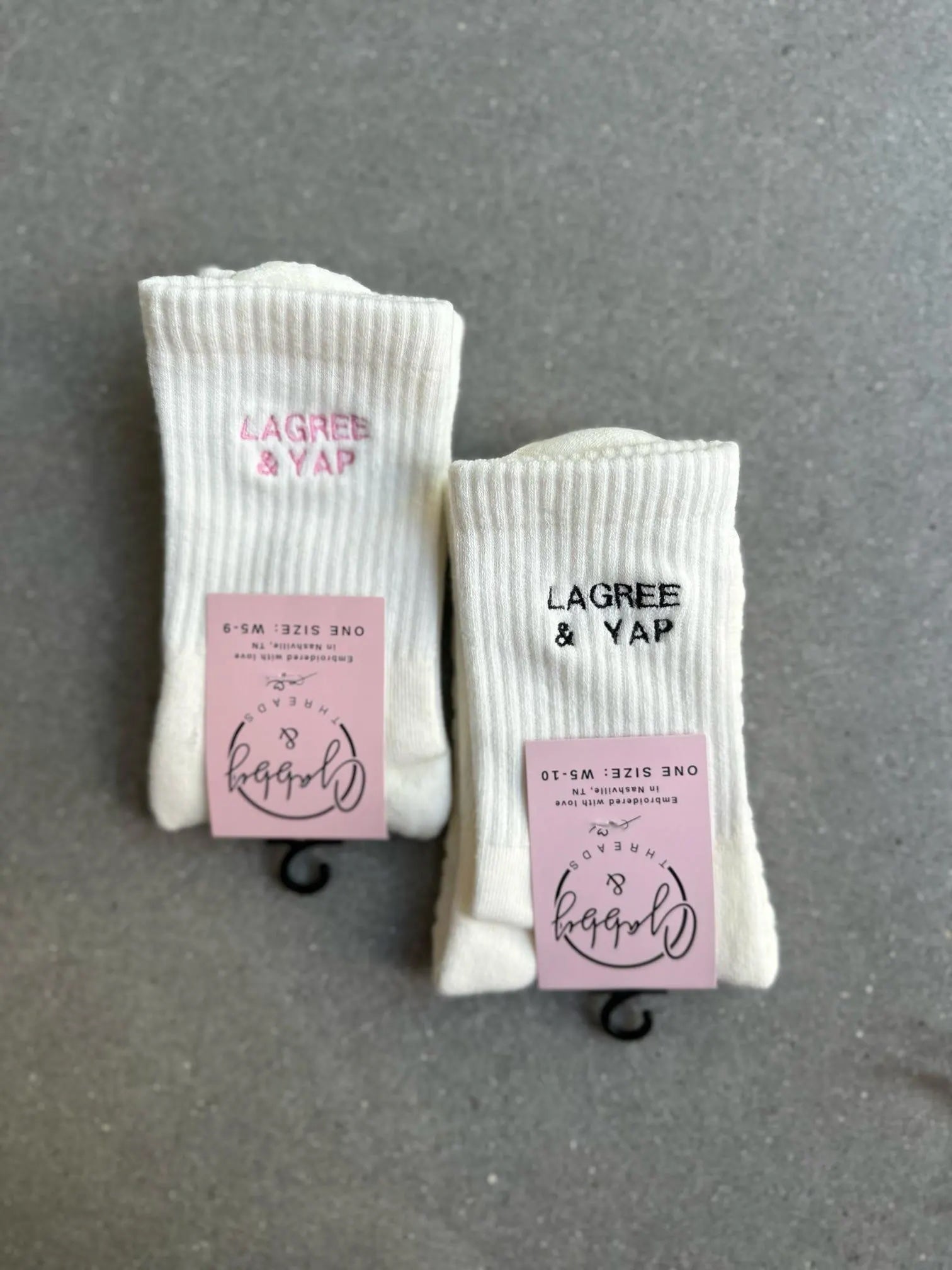 two pairs of white grip socks with the words "lagree and yap" in pink and black thread, lay on a concrete floor