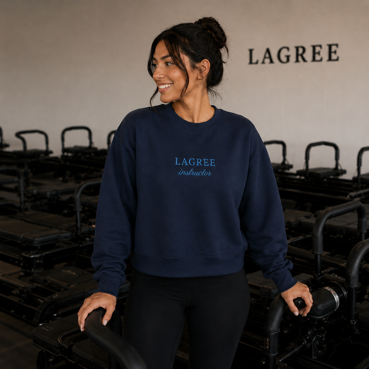 a woman is in a lagree studio wearing an embroidered crewneck that has the words "lagree instructor" embroidered on it
