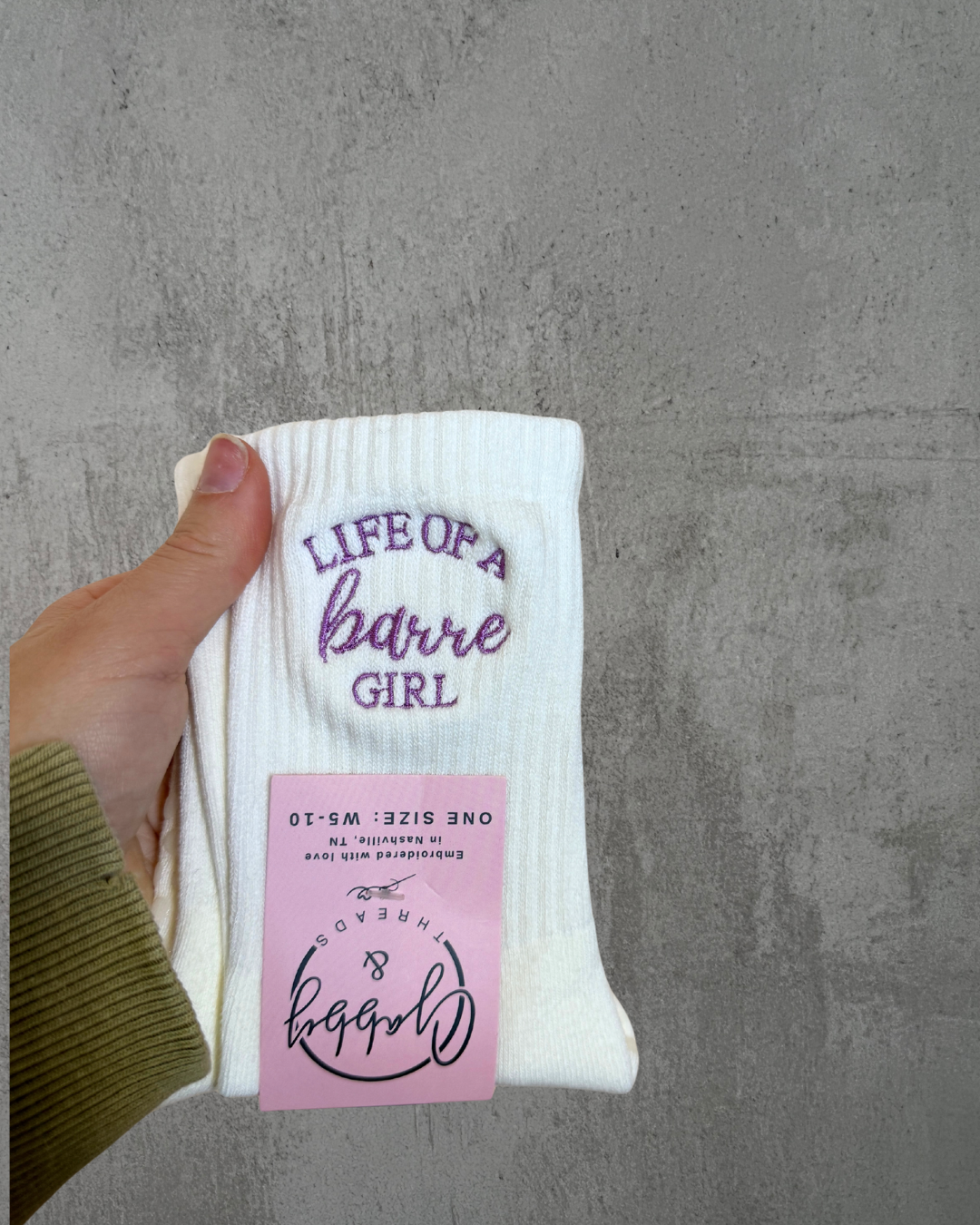 a pair of white grip socks against a gray background. The white grip socks have the words "life of a barre girl" embroidered on them in lilac thread