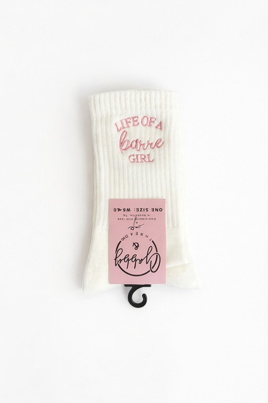 a pair of white grip socks against a white background. The white grip socks have the words "life of a barre girl" embroidered on them in pink thread