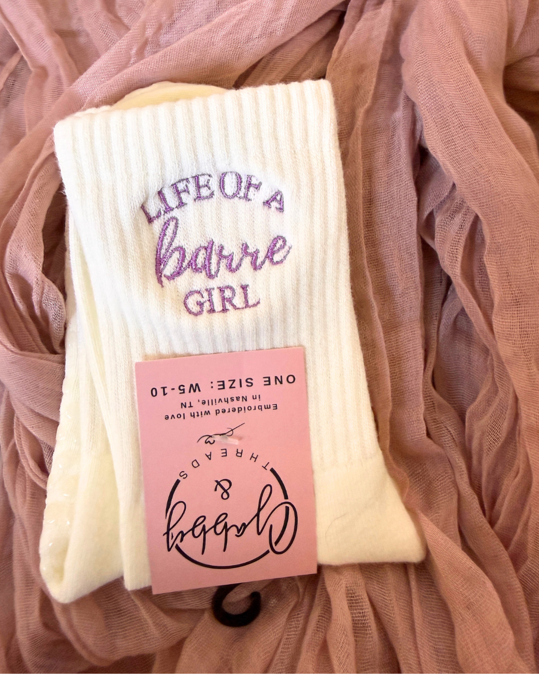 a pair of white grip socks against a pink background. The white grip socks have the words "life of a barre girl" embroidered on them in lilac thread