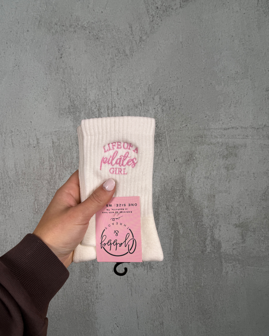 a hand holds up a pair of white grip socks with the text "life of a pilates girl" embroidered on it against a grey background
