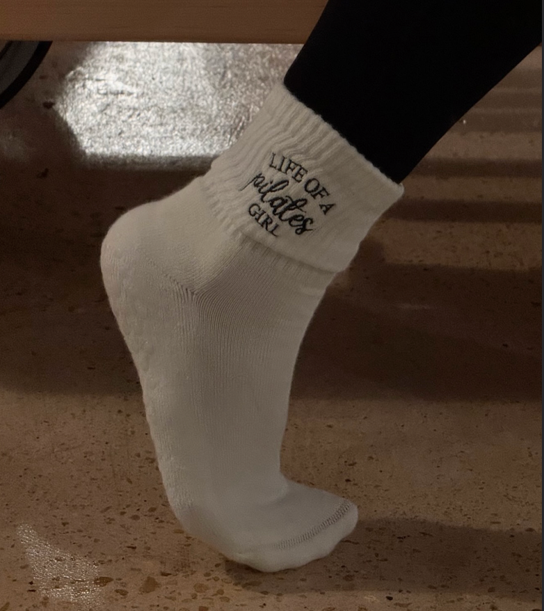 a person is wearing a pair of white grip socks next to a reformer that say "life of a pilates girl" 
