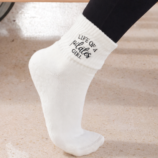 a person is wearing a pair of white grip socks next to a reformer that say "life of a pilates girl" 