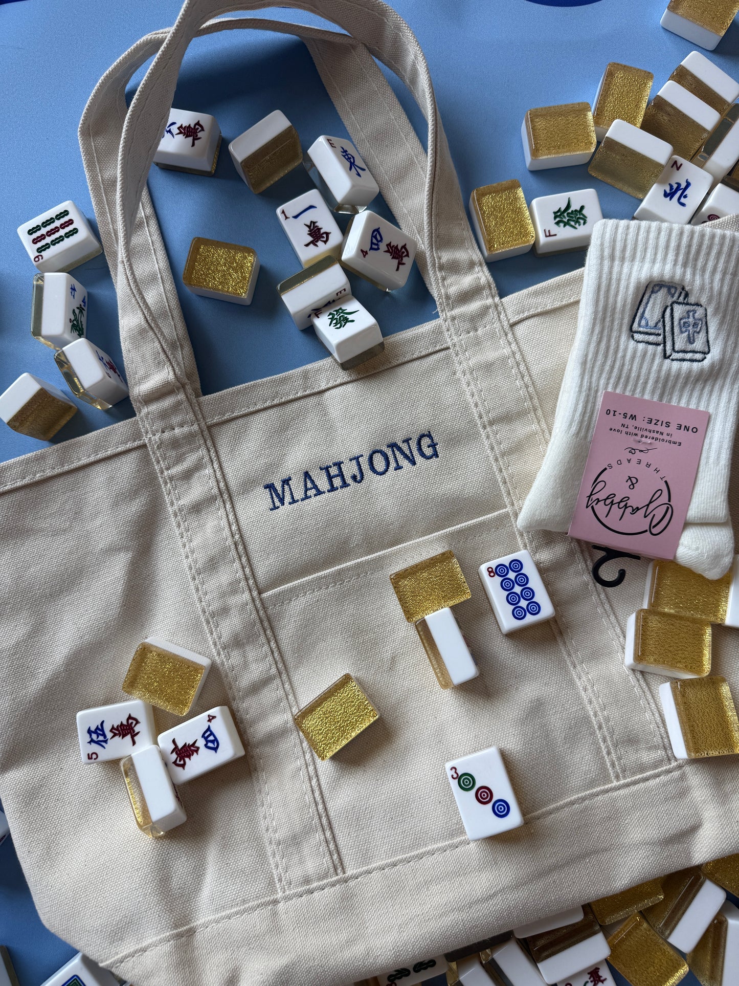 A beige canvas tote bag with 'MAHJONG' printed in navy blue on the front pocket, styled with miniature mahjong tiles and a pink card on a blue background.