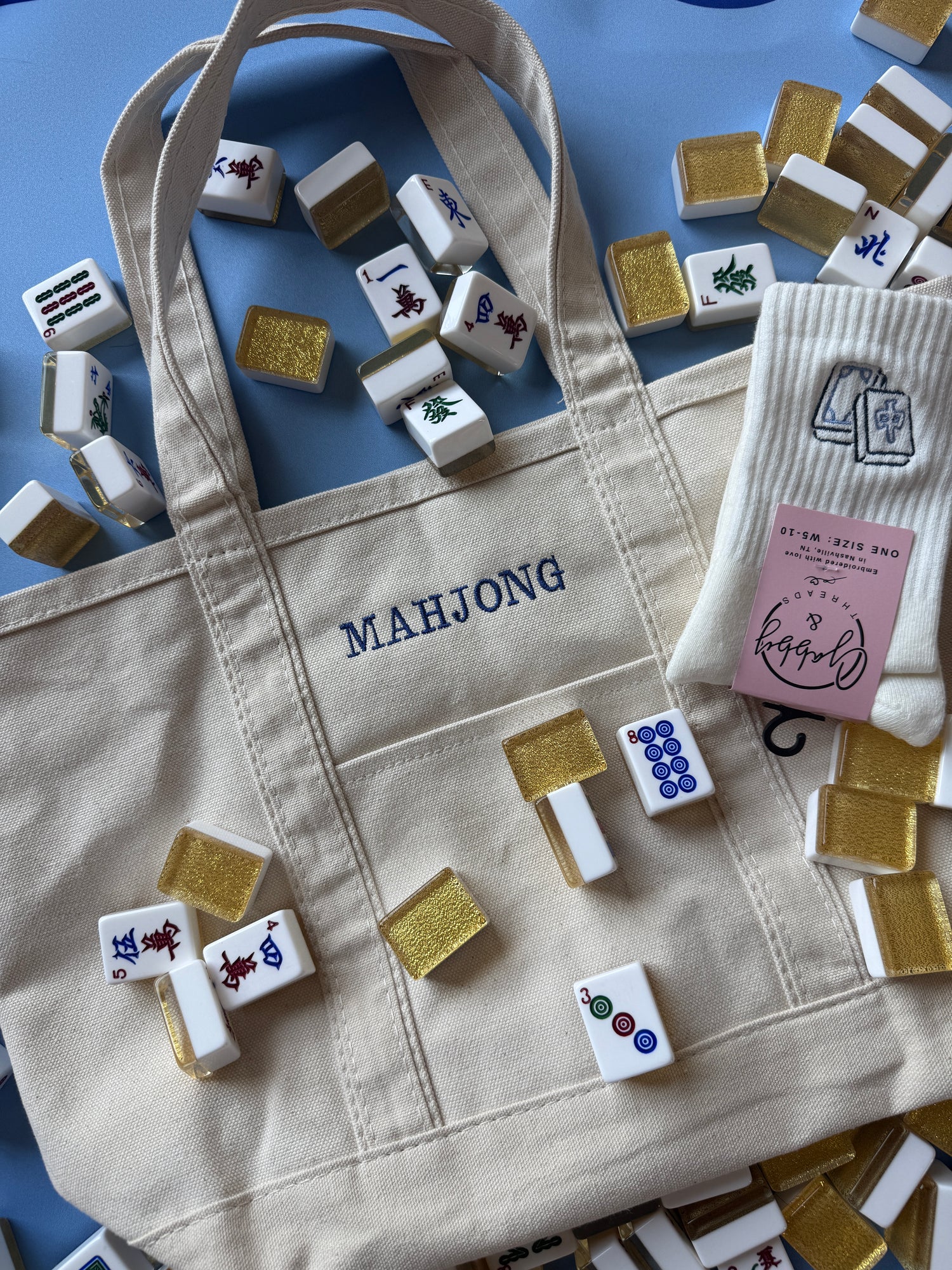 A beige canvas tote bag with 'MAHJONG' printed in navy blue on the front pocket, styled with miniature mahjong tiles and a pink card on a blue background.