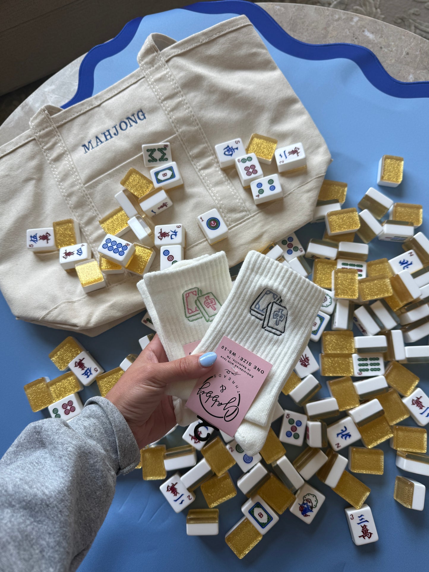 A hand holds a pair of white ribbed socks with pink tags, surrounded by scattered mahjong tiles and a cream canvas tote bag printed with 'MAHJONG' on a blue surface.