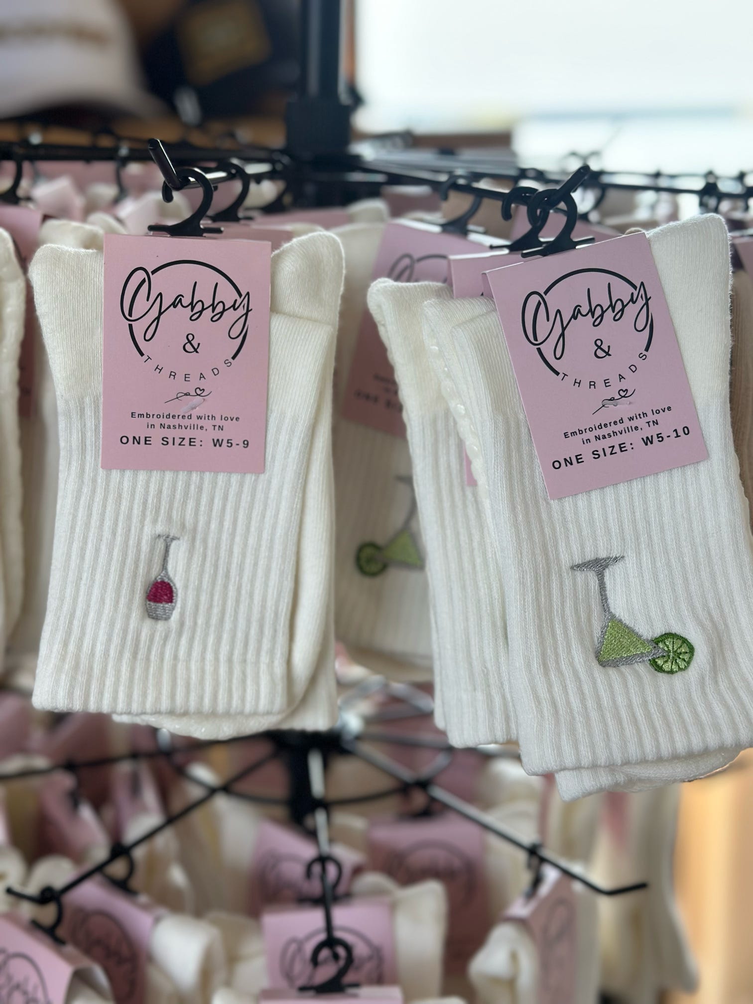 Two pairs of white grip socks hanging on a retail rack. One pair has an embroidered margarita and the other has an embroidered wine glass