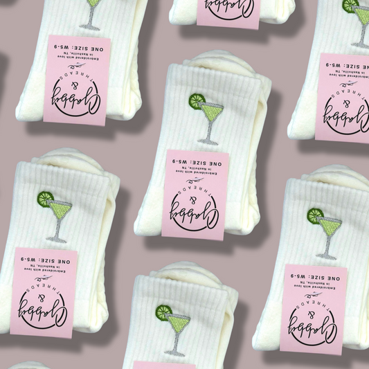 White socks with pink labels and embroidered green margaritas on a mauve gray background